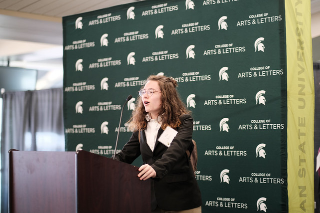 Woman with brown wearing a black blazer is standing behind a podium and speaking into a microphone 