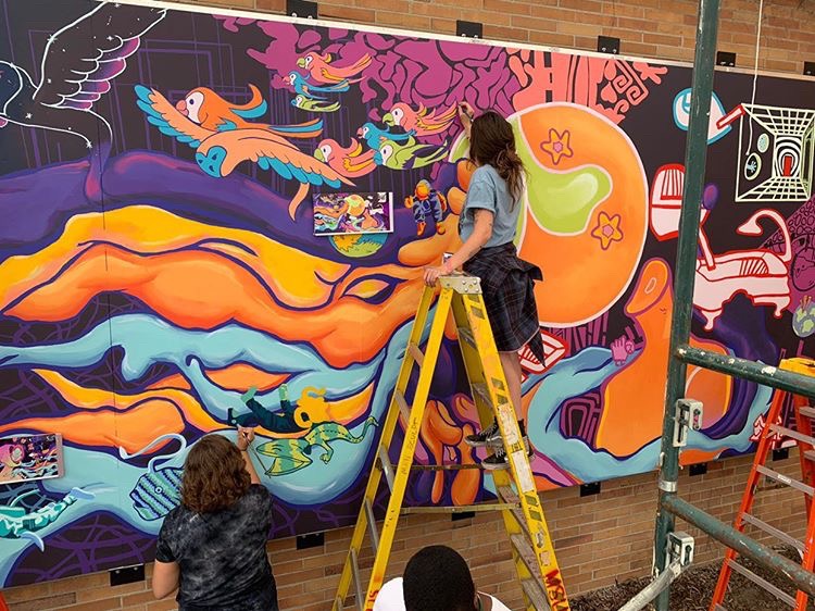 Two women painting a mural. One in standing on a ladder and the other is standing on the ground painting