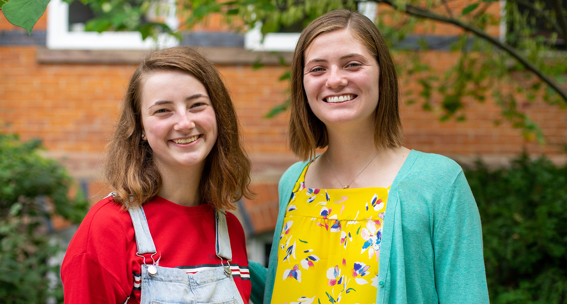 Two girls with short brown hair standing next to each other smiling in front of a brown brick building and green trees