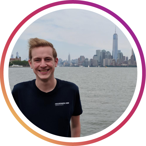 a man with blonde hair standing by a body of water with a city behind him