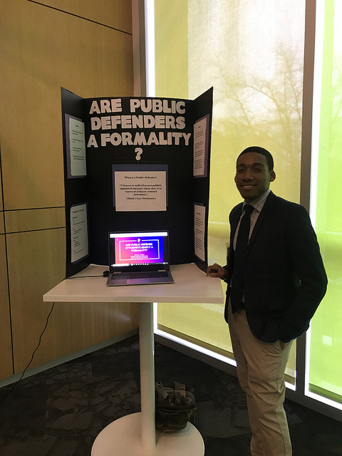 a man in a suit and tie with a poster board and laptop giving a presentation