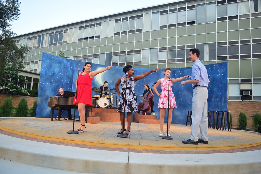 three singers singing and pointing at an actor standing to their right, a band is playing instruments in the background