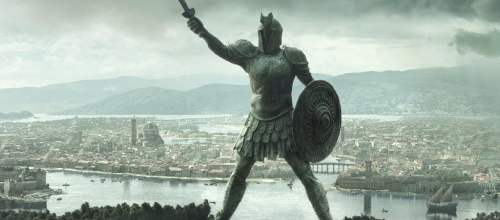 a large warrior statues stands with a shield and sword. Behind it is a large city near the water.