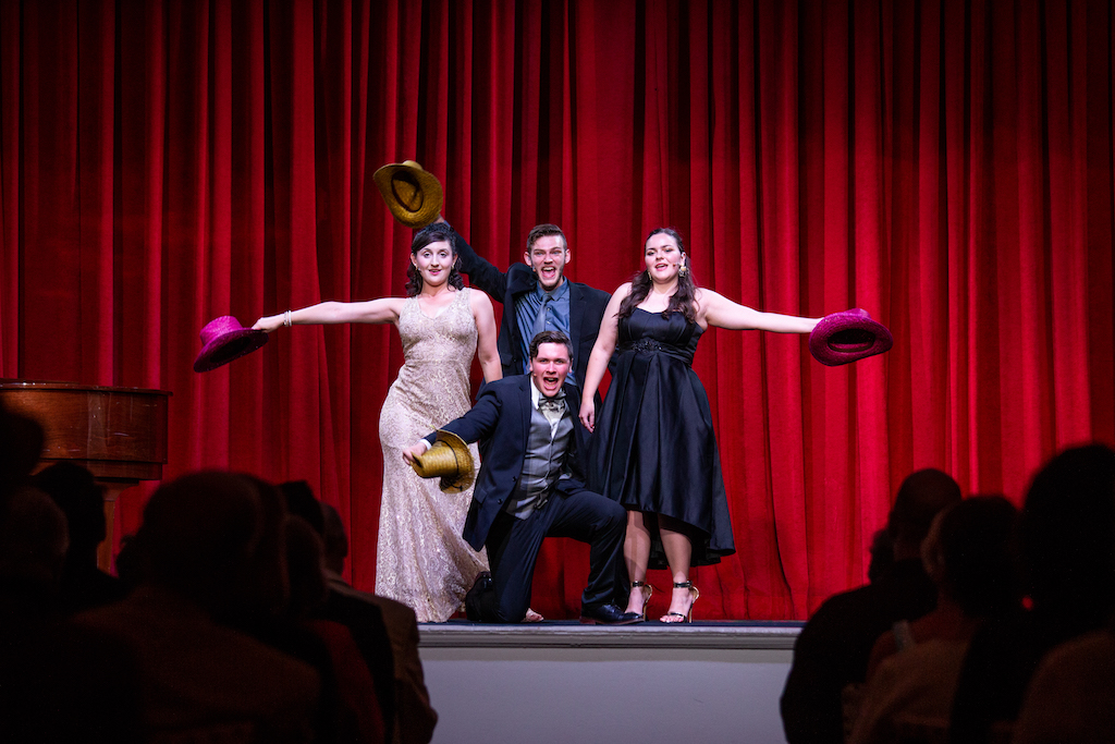 group of four people who are dressed in formal attire and are holding colorful hats in the air in front of a red curtain