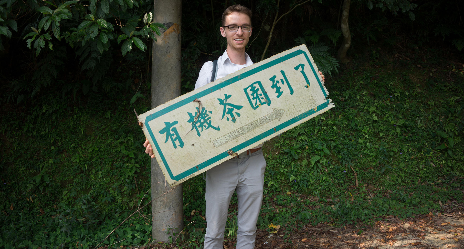 Man wearing gray pants holding a sign in front of greenery