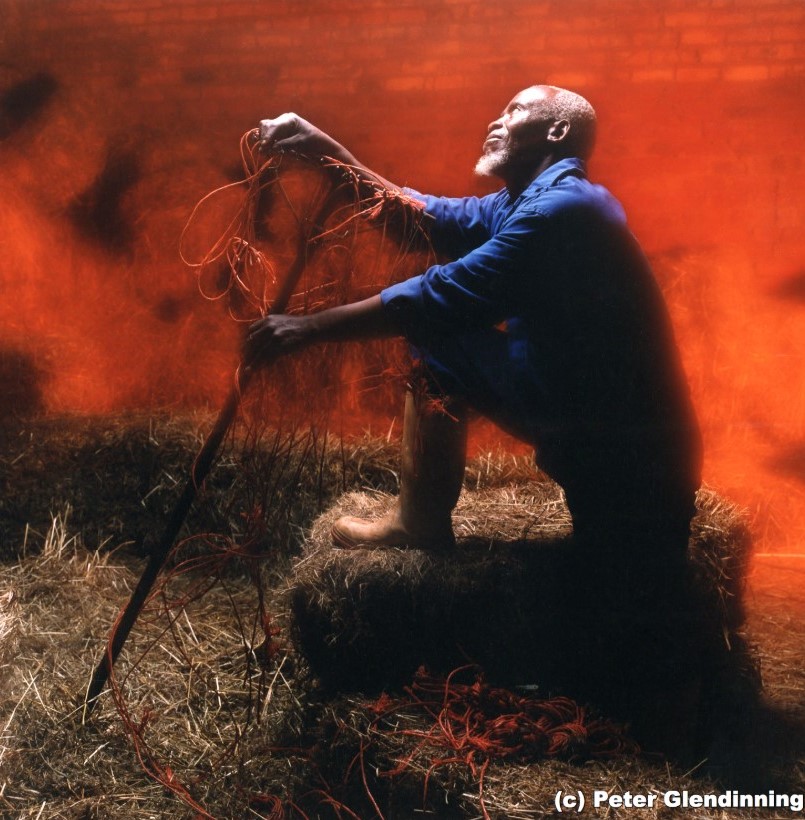 Portrait of a man with one leg up on a stack of hay holding a rake in front of a red background with brick