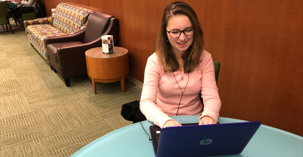 a young woman sitting and using her laptop with earbuds in. she is wearing large glasses and a pink long-sleeved top