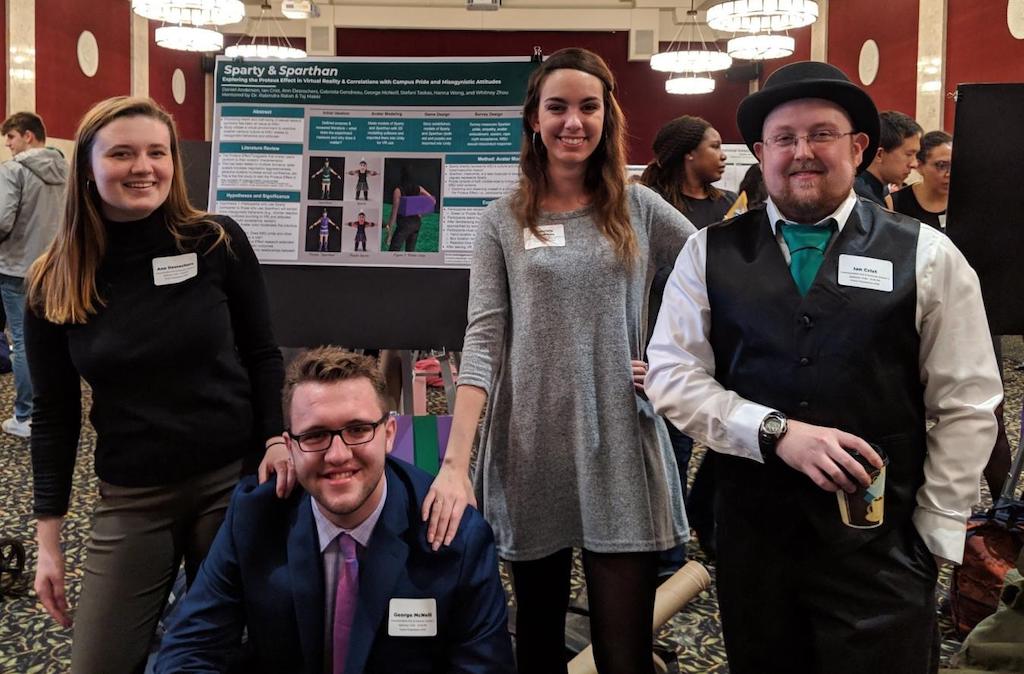 four adults standing around a poster and smiling at the camera