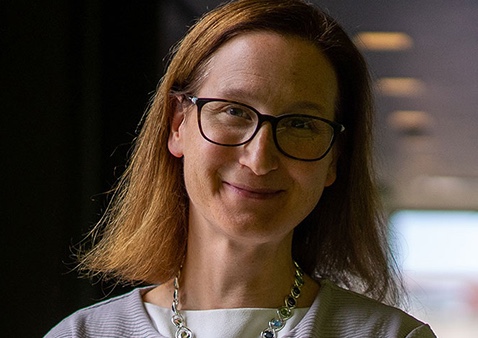 a women with short hair wearing glasses, a necklace, a white top with a grey jacket