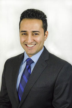 portrait of a smiling man with short, slicked back black hair. He is wearing a suit with a light blue button-up and dark blue tie