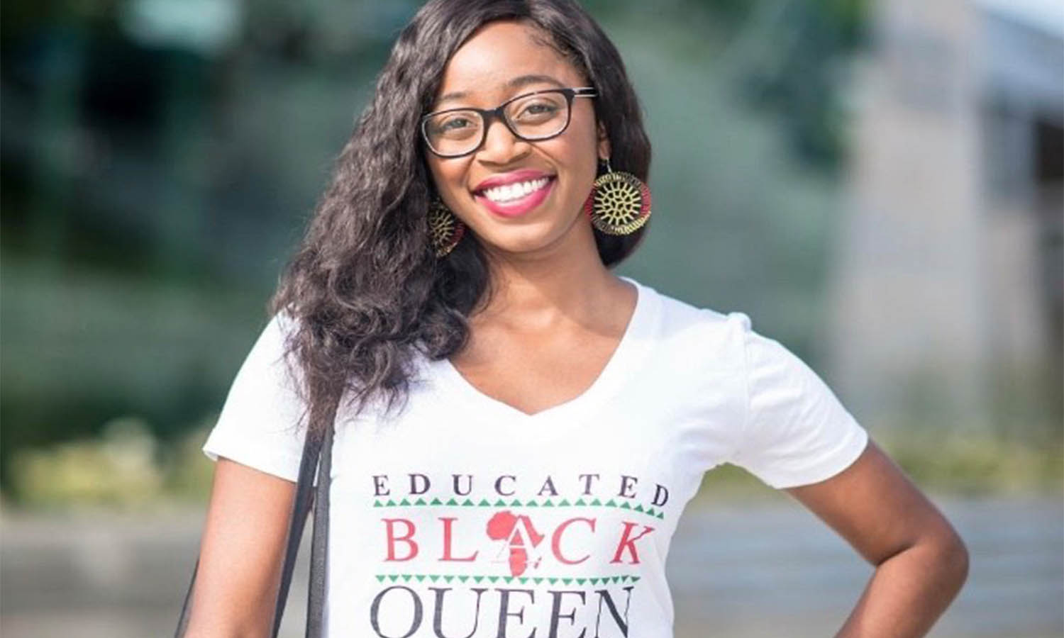 Woman with black hair is smiling. She is wearing black-rimmed glasses, circular earrings, and a white v-neck shirt that says
