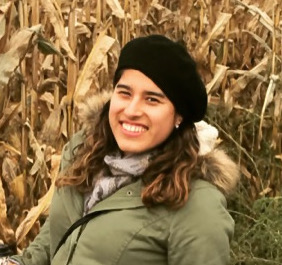 headshot of a woman wearing a black hat with long brown hair and an olive green coat