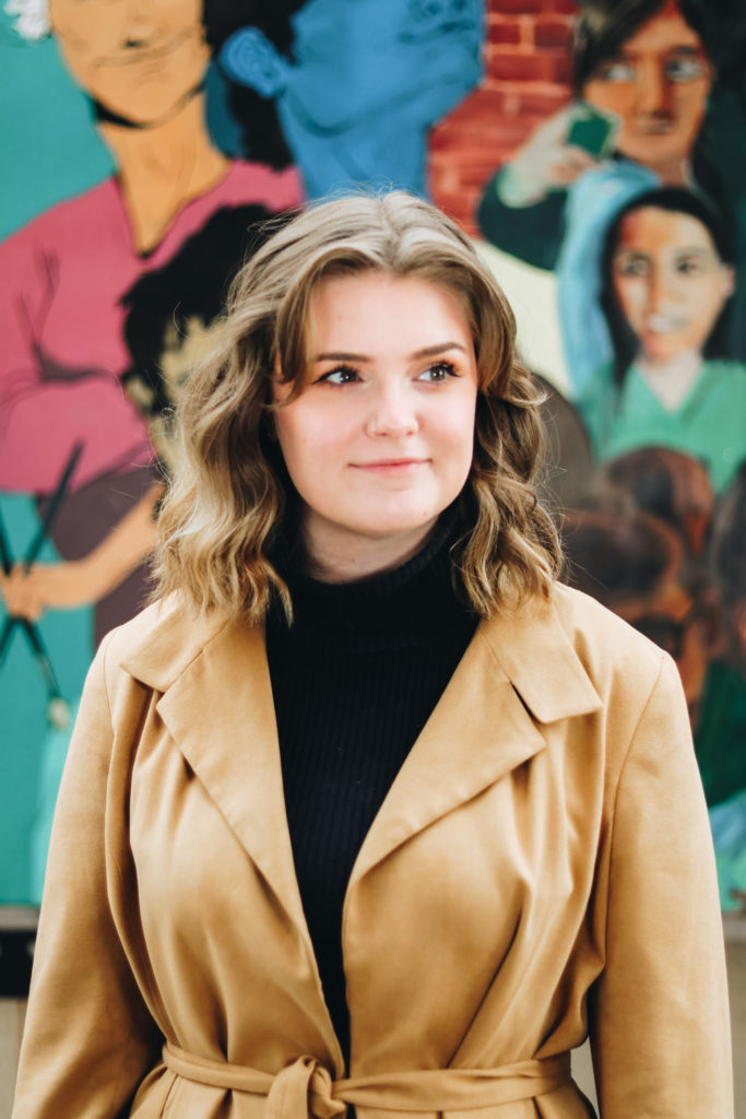 Photo of a woman with shoulder-length wavy brown hair. She is looking to the side of the camera and is wearing a tan jacket and black sweater. Behind her is a mural painting of a group of people.