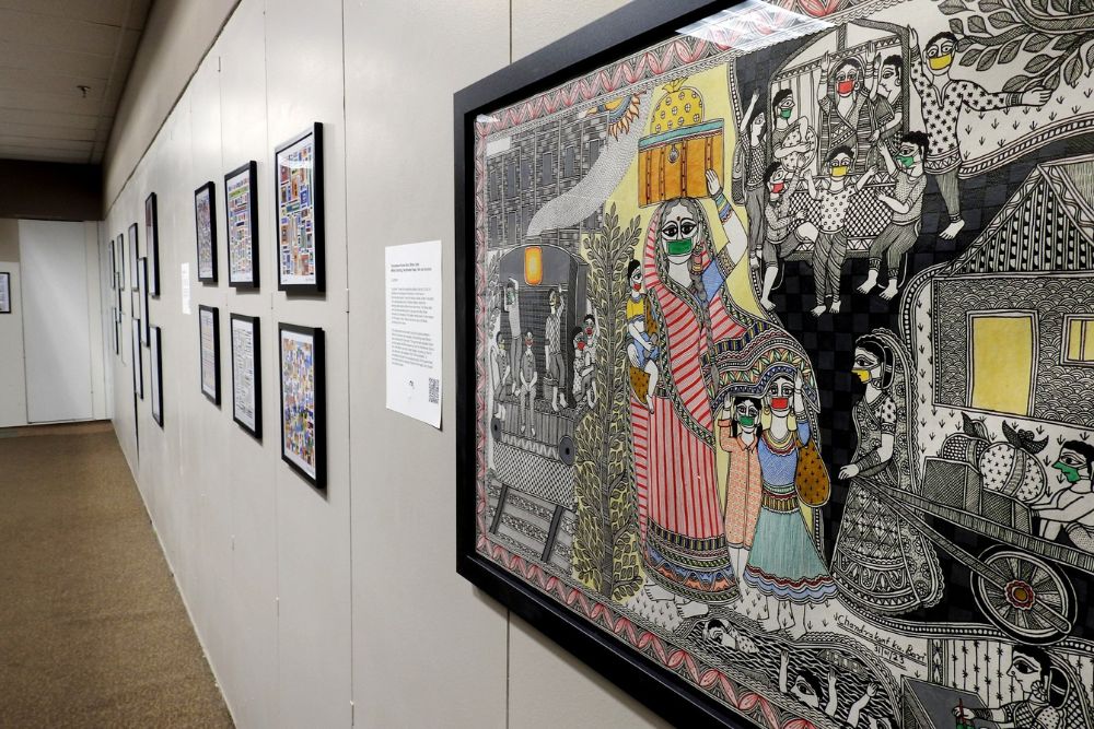 A framed illustration depicting Indian men and women with COVID-19 face masks hangs on the wall of an exhibit. An Indian woman holding her young son and balancing a box on her head is centered while her two young daughters stand beside her.