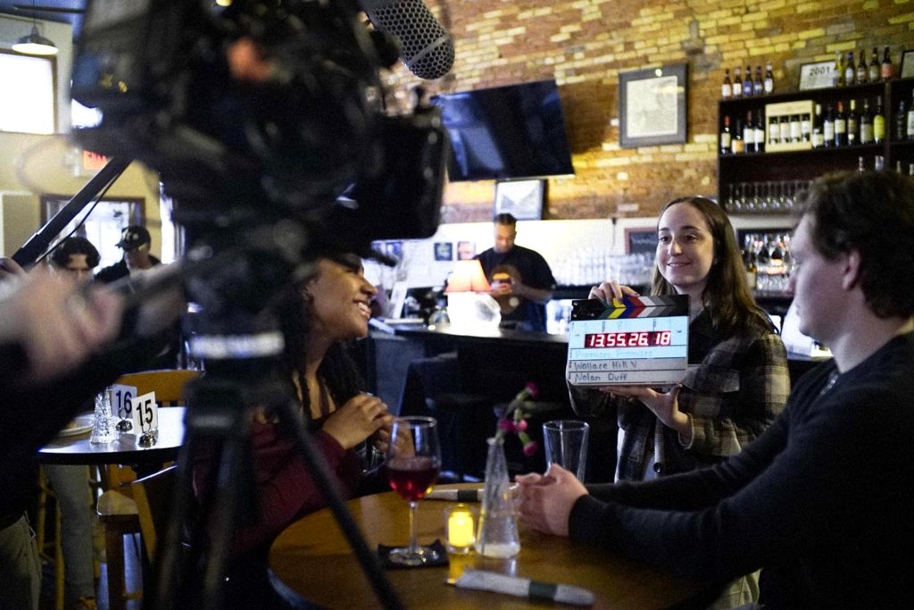 A camera in the foreground stands in front of a table with two students behind it at restaurant. A smiling student in front of the table holds a clapperboard and looks into the camera.