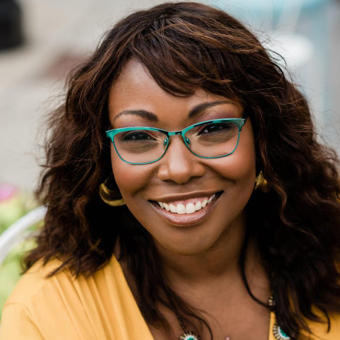 MSU Associate Professor Kinitra Brooks smiles wearing a yellow dress and glasses.