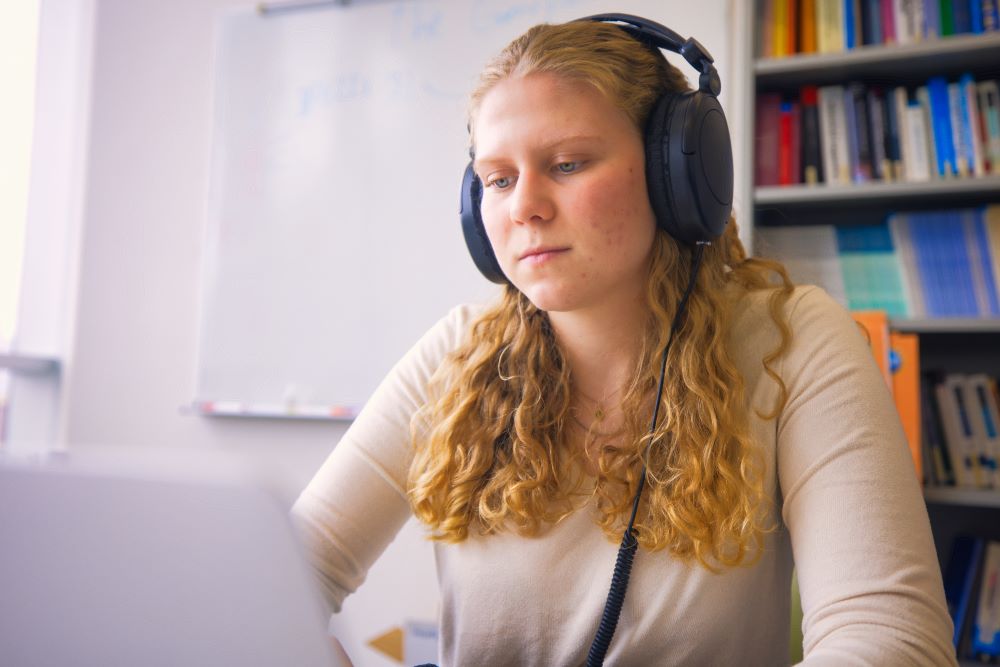 A person with headphones and curly hair works on a laptop while sitting at a table.