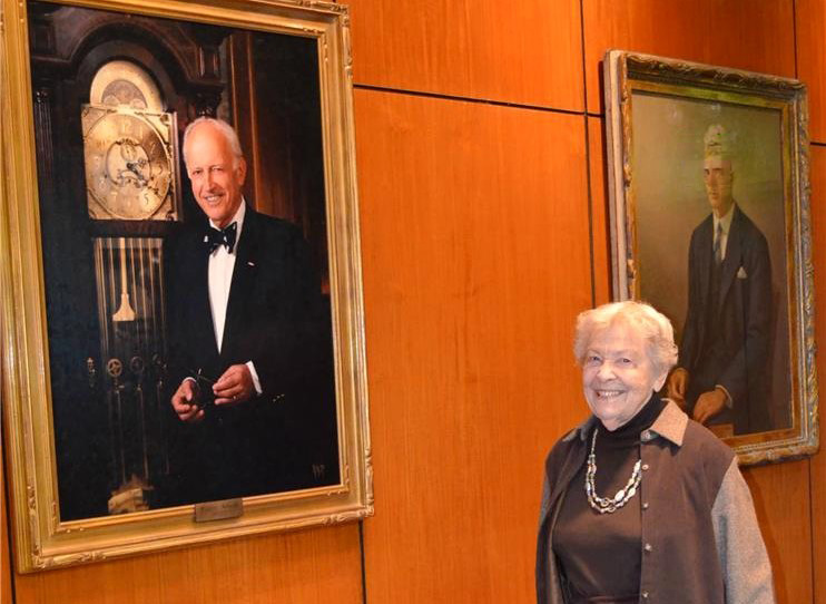 Pauline, a woman wearing a brown shirt and white necklace, stands next to her husband's portrait.