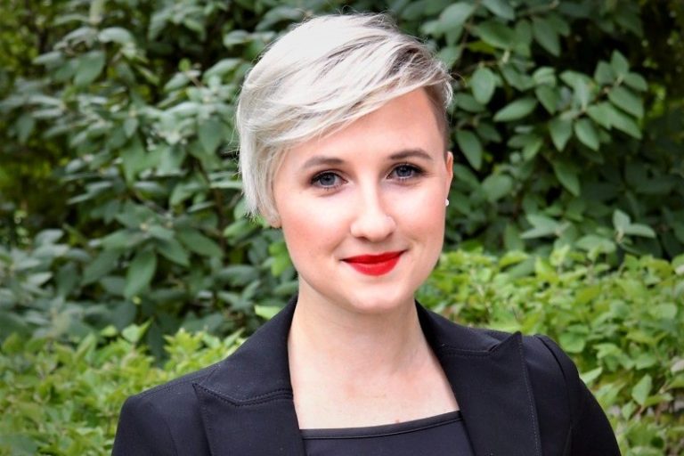 A picture of a woman with short blonde hair in a black suit standing in front of a leafy background.