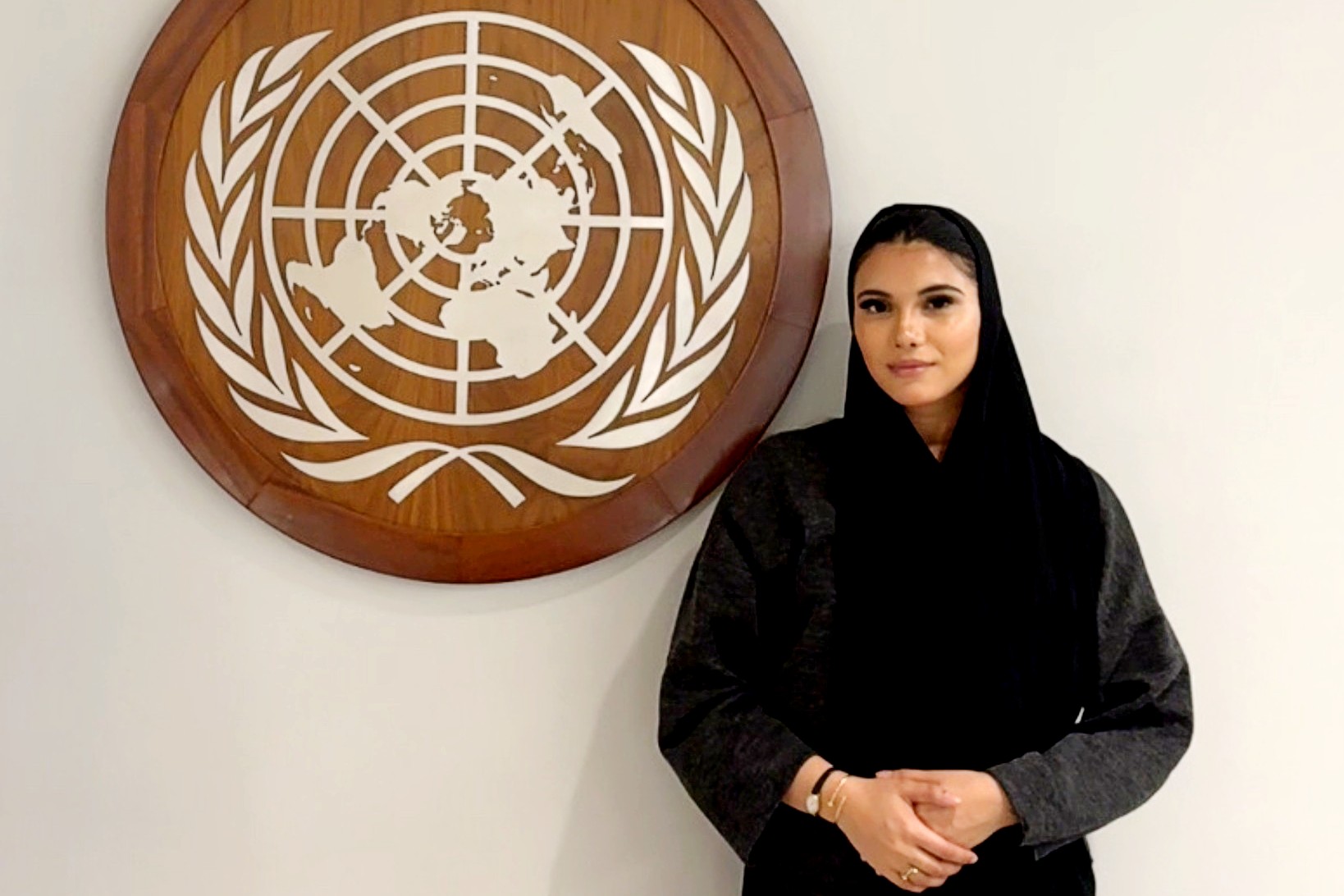 A picture of a woman in all black with gold rings and a watch standing next to a wooden icon with leaves and a map of the world.