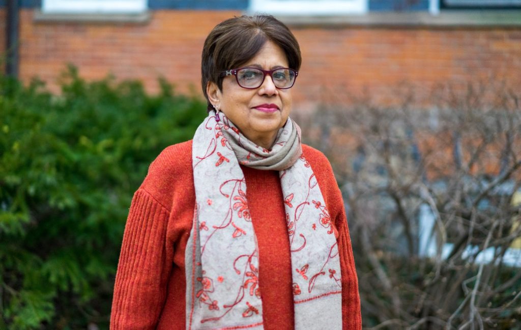  A woman with short dark hair and glasses stands outside in front of a brick wall and greenery. She is wearing an orange sweater and a beige scarf with an embroidered floral pattern.