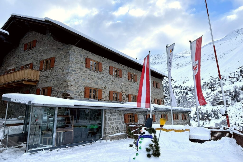 The University of Innsbruck's Alpine Retreat Center - the photo shows a three-story stone building with several small windows with brown shutters. Outside there are three long banners and a snowman. The building  is nestled in a mountain with the top of the mountain that can be seen in the background.