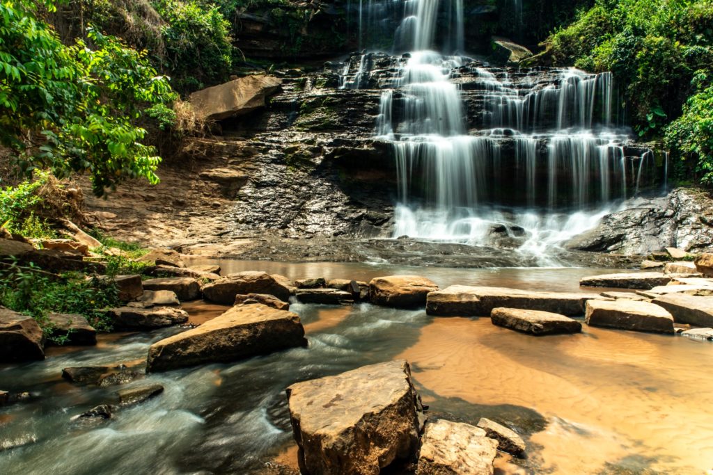 A scenic view of a cascading waterfall surrounded by lush green foliage. The water flows over dark rock formations, with a clear stream running through a foreground of smooth stones