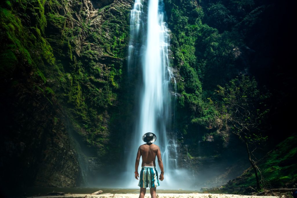 A man wearing swim trunks and a wide-brimmed hat stands at the base of a towering waterfall. The cascading water is framed by lush green cliffs.