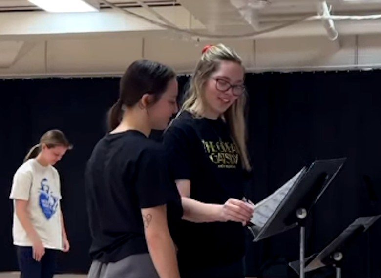 Two performers stand close together, reviewing sheet music on a black music stand.