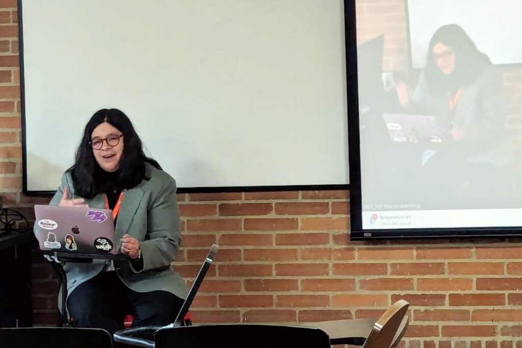 A woman with long dark hair and glasses is seated in front of a laptop covered in stickers, speaking to an audience. She wears a gray blazer and an orange lanyard. Behind her, a projection screen displays a live video of her presentation. The setting has exposed brick walls and classroom seating.