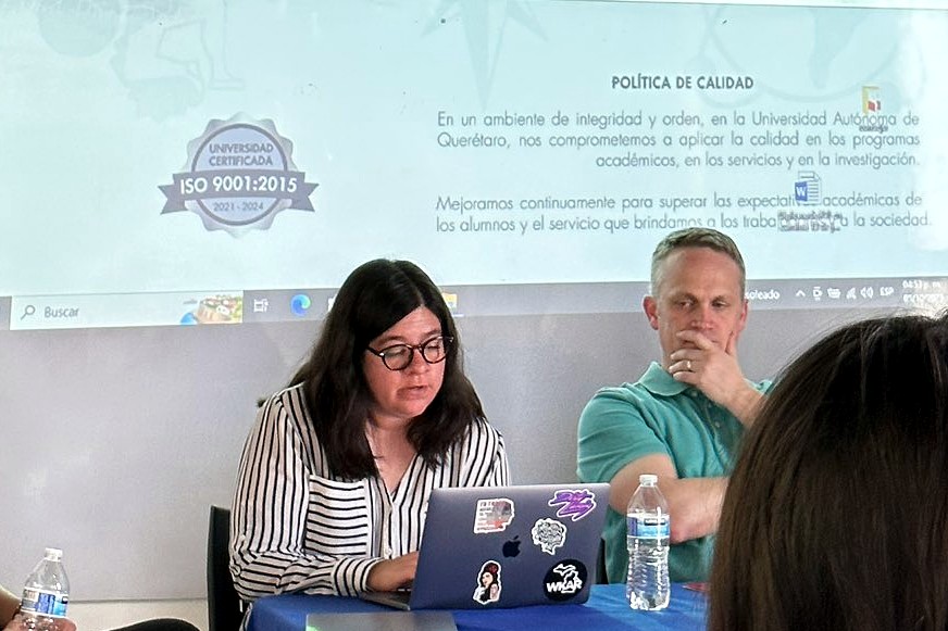 A woman with long dark hair and glasses sits at a table with a laptop covered in stickers, concentrating as she types. Beside her, a man in a teal polo shirt sits with his hand on his chin, appearing thoughtful. Behind them, a projected slide displays Spanish text about academic quality policies.