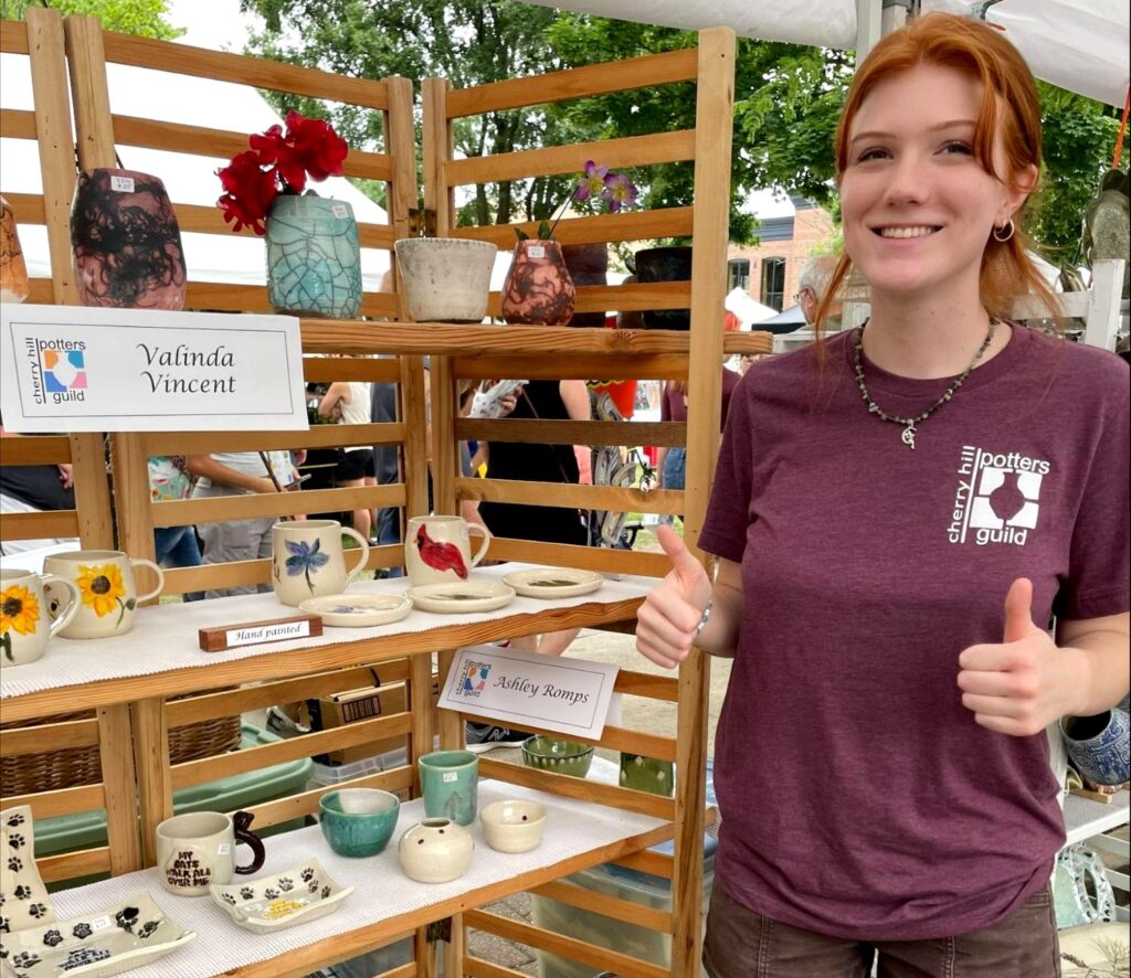 Woman standing next to a shelf that contains pottery that is for sale. The woman has red hair that is pulled up into a pony tail and is wearing small gold hoop earrings and a burgundy-colored T-shirt that says "Cherry Hill Potters Guild" on it. The woman is giving two thumbs up. And the shelf includes the names of the potters. One being 'Valinda Vincent" and the other being 'Ashley Romps" who also is the person in the photo. 