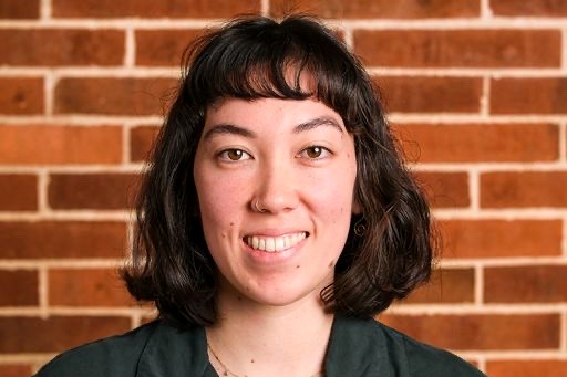 Head shot of a woman with shoulder-length dark brown hair and short bangs. She is wearing a small gold nose ring and a smile. 