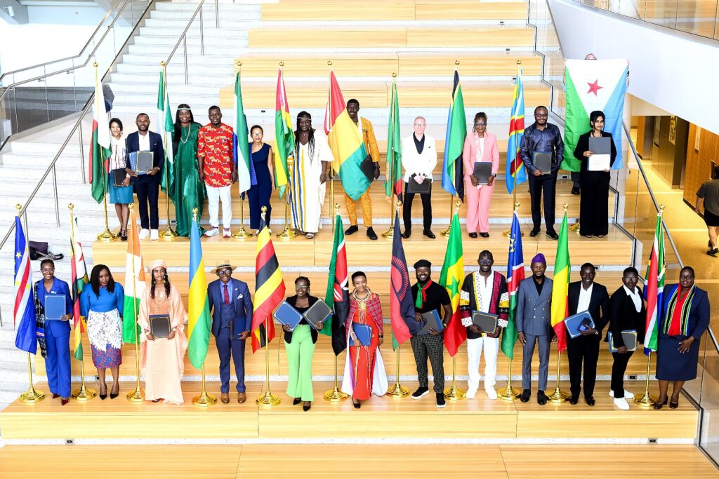 A group of people stand on wide wooden steps inside a bright, modern building. They are arranged in two rows, with national flags from various African countries positioned between them.