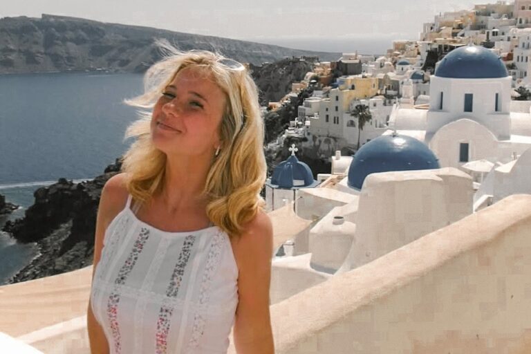 woman with long blond hair standing with a Greek village in the background and a large body of water.