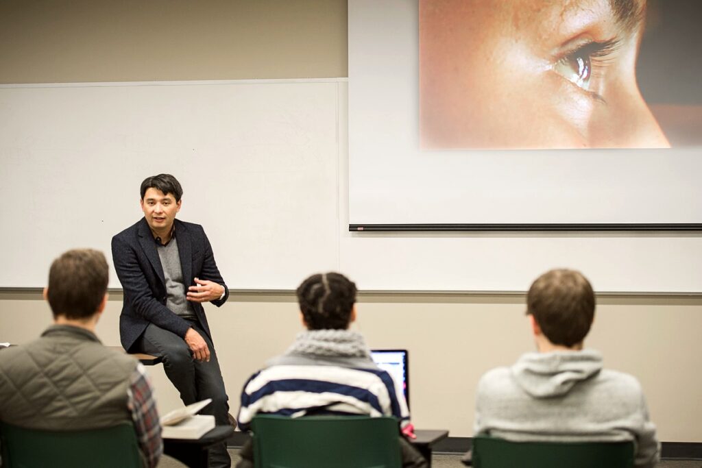 Professor Joshua Yumibe sits on a desk at the front of a classroom, engaged in conversation with students. He faces the class, gesturing as he speaks. Behind him is a whiteboard and a large projection of a close-up image of an eye, partially visible. Three students are seated in the foreground, listening attentively.