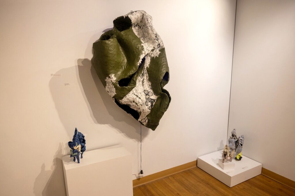 A large abstract wall-mounted sculpture in green and white dominates the corner of the gallery. Two smaller colorful ceramic sculptures stand on white pedestals nearby, their rough textures contrasting with the smooth gallery walls.