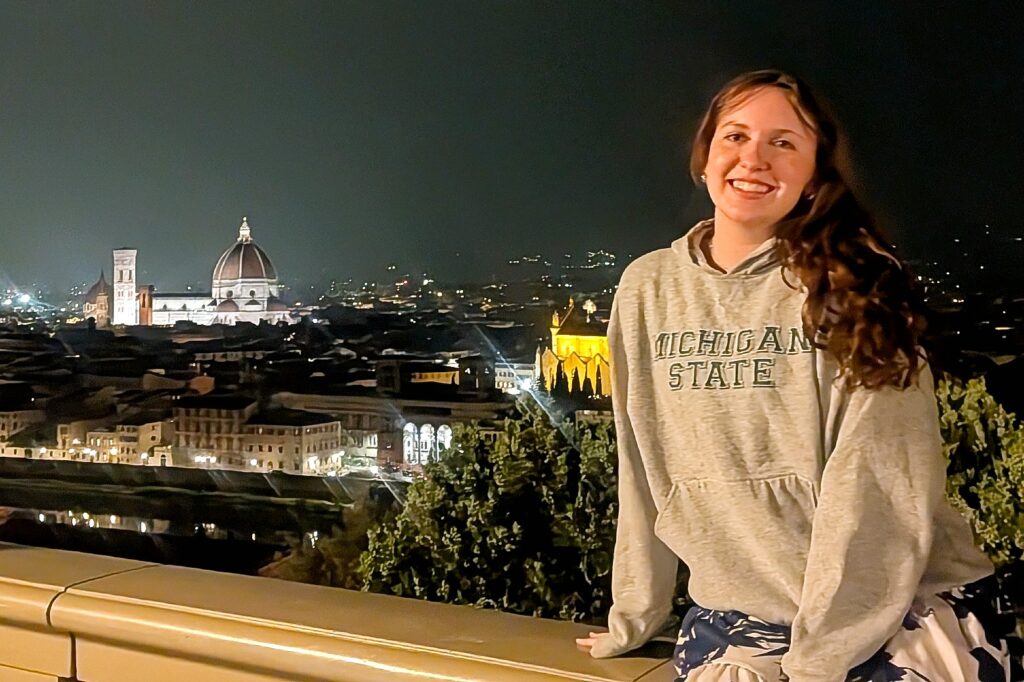 A student wearing a gray Michigan State University hoodie smiles at night with the illuminated skyline of Florence, Italy, behind her. The Florence Cathedral, or Duomo, with its iconic dome and bell tower, is clearly visible in the background.