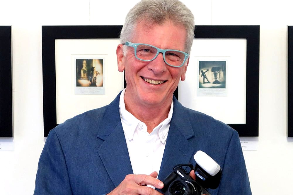 Portrait of an older man with light hair and glasses, smiling while holding a Canon camera with a flash attachment. Framed photographs hang on the wall behind him in a gallery setting.