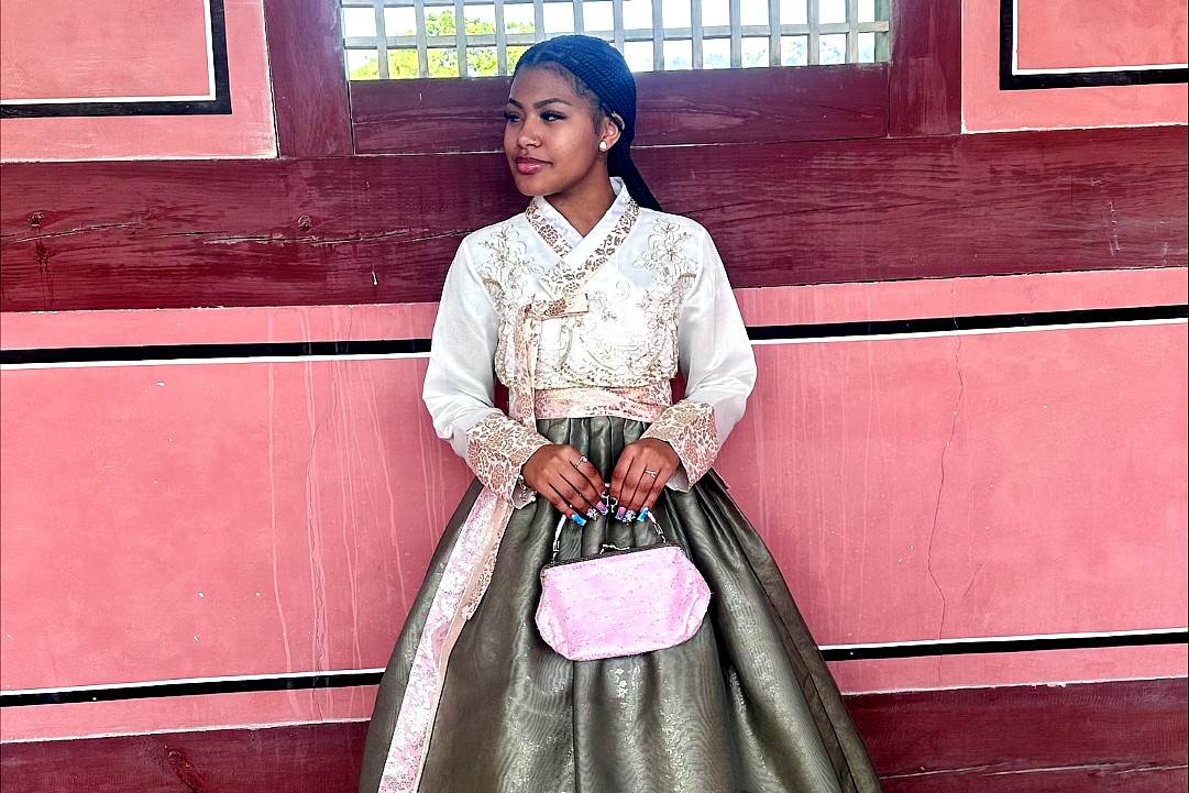 A woman stands against a pink and red traditional Korean building wall, wearing a hanbok with a white embroidered top, olive green skirt, and pale pink trim, holding a small pink handbag.