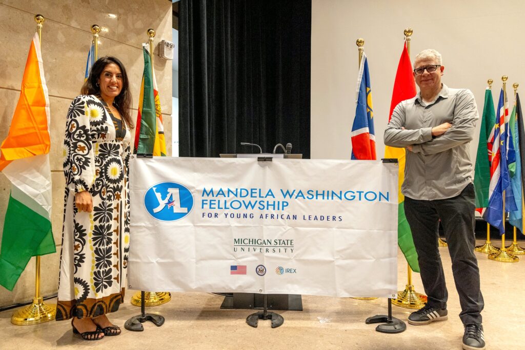 Two people stand on either side of a banner for the Mandela Washington Fellowship for Young African Leaders at Michigan State University. The woman on the left wears a long, patterned dress and smiles at the camera. The man on the right, wearing glasses, a gray shirt, and dark pants, stands with arms crossed.