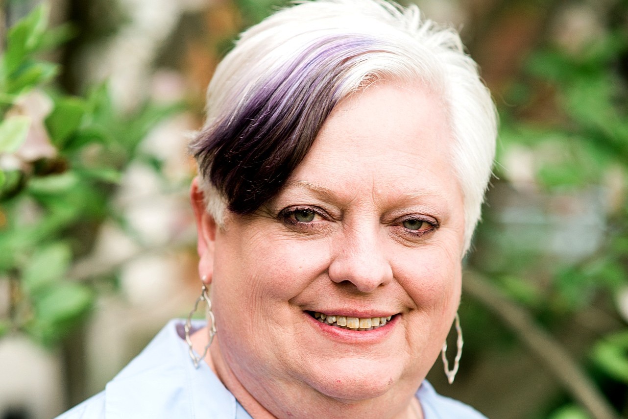 A close-up outdoor portrait of a person with short, white hair with a purple streak, wearing dangling earrings and a light-colored shirt, smiling at the camera with greenery in the background.
