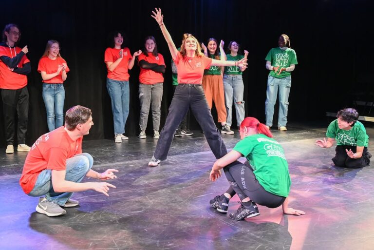A group of college-aged performers on a theater stage, energetically acting out a scene. One performer stands center stage with arms outstretched, surrounded by others wearing red and green shirts, some crouching and others clapping or laughing in the background.