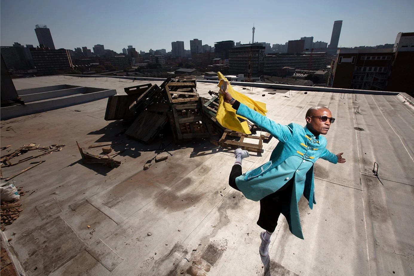 A man in a bright turquoise coat and sunglasses strikes a dynamic, mid-leap pose on a city rooftop, with a pile of broken wooden pallets and a yellow cloth behind him. The city skyline stretches across the background under a clear sky.