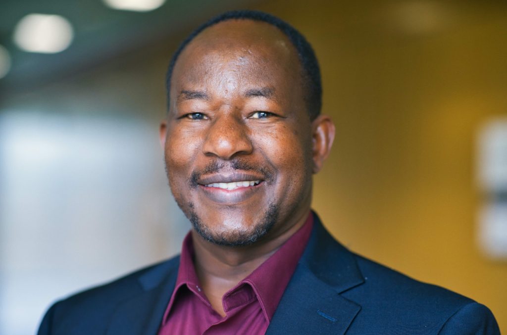 Headshot of Black man smiling, wearing dark blue suit and maroon shirt.