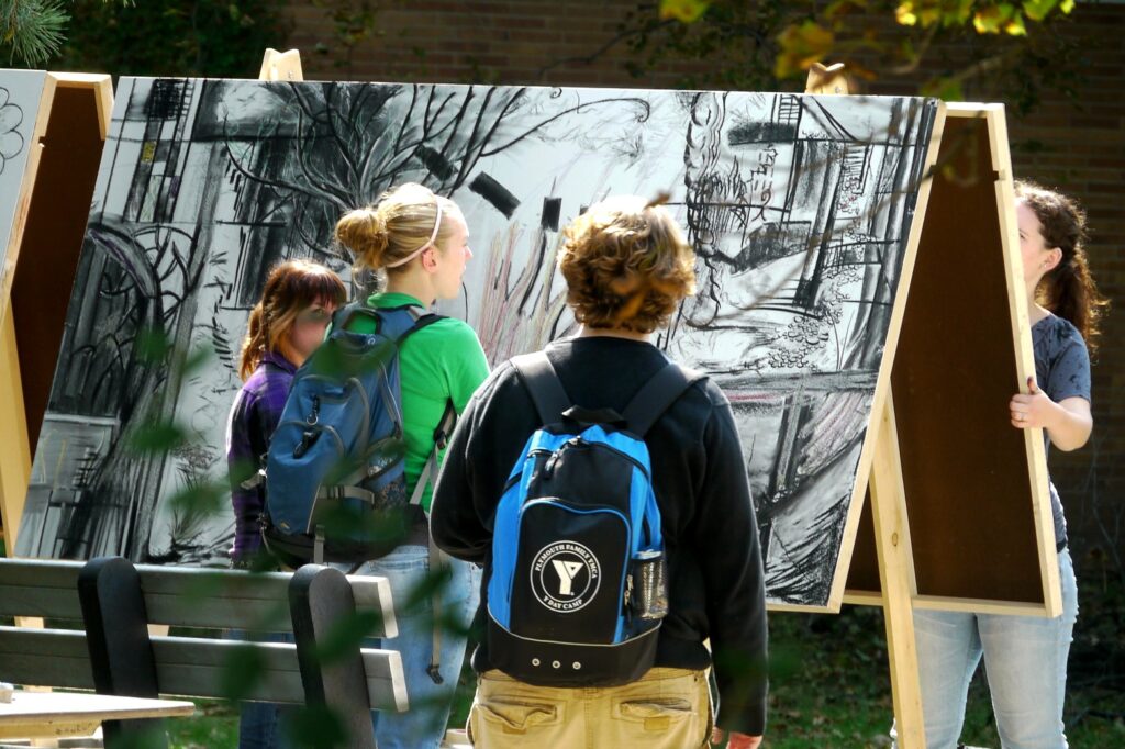 A small group of students gathers around a large easel with a charcoal drawing of trees and abstract shapes, with another student on the side of the frame.