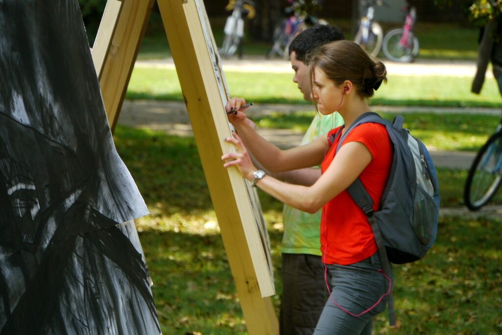 Two students with backpacks draw on tall easels set up outdoors; one wears a bright orange shirt and gray pants, while the other, slightly behind, focuses on sketching in the background