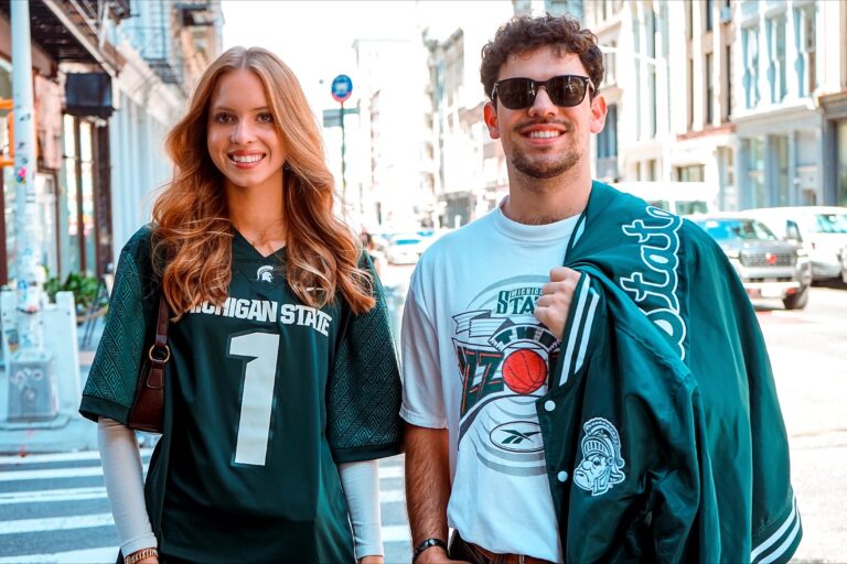 Two students stand side by side on a city street. One wears a dark green Michigan State jersey, and the other wears a white shirt with a green MSU jacket draped over his shoulder. Both smile at the camera.