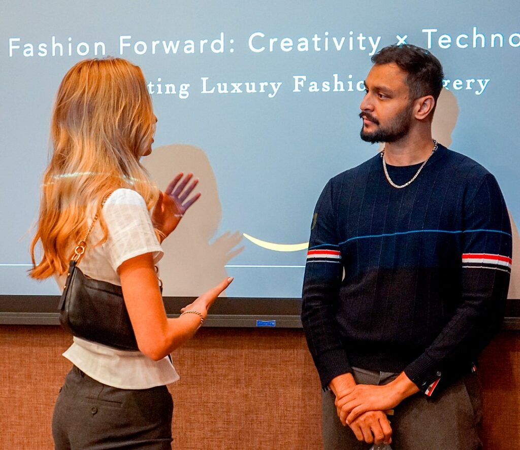 Woman with long blonde hair talking to a man with black hair and a beard. The woman has her hands raised and is gesturing while she speaks. The man is looking at her. On the screen behnd them it says "Fashion Forward: Creativity x Technology.