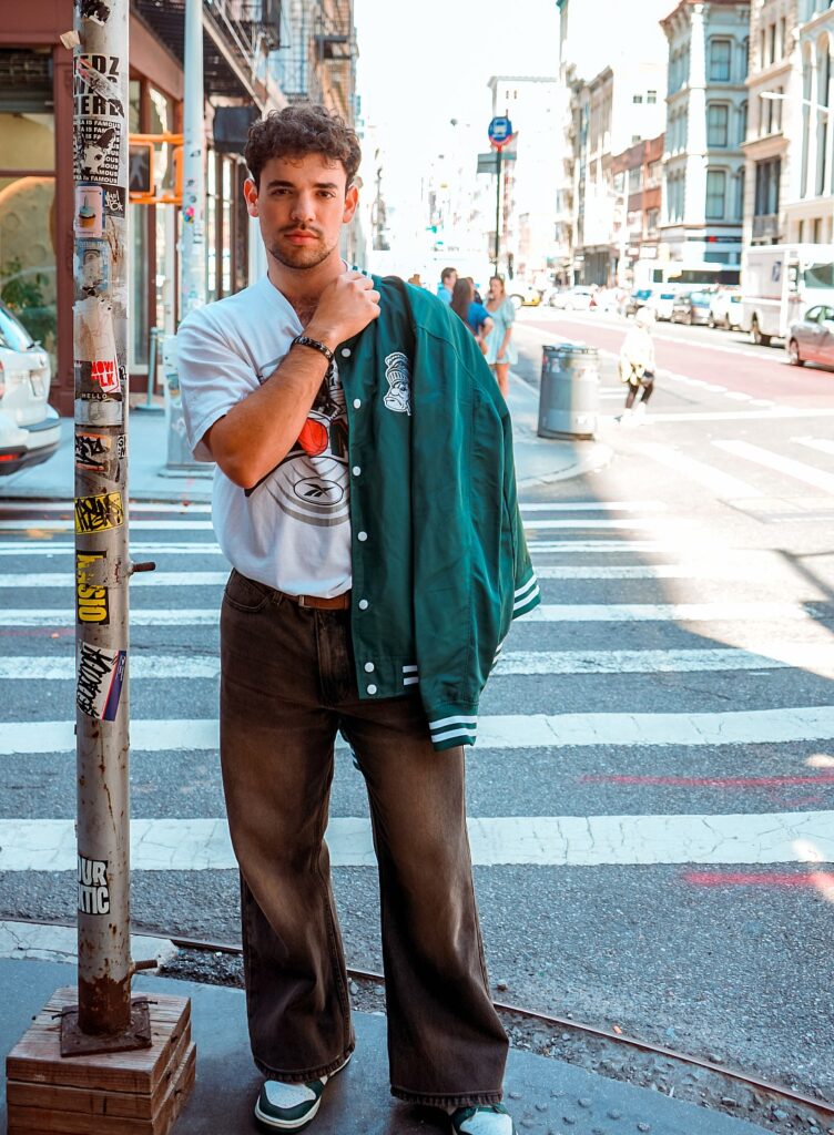 A student wearing a white T-shirt and dark pants poses with a green Michigan State jacket over his shoulder.